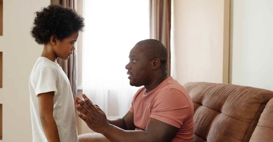 A father talking to his son. The father is sitting on the chair and has his arms out as he is talking