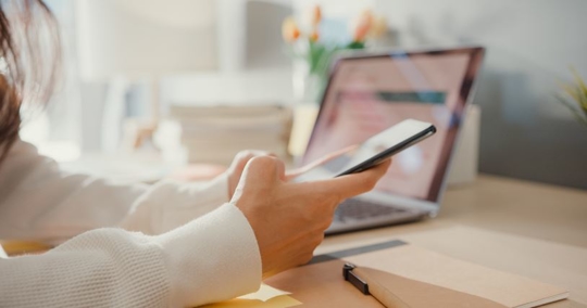 Person holding their smartphone with laptop in background